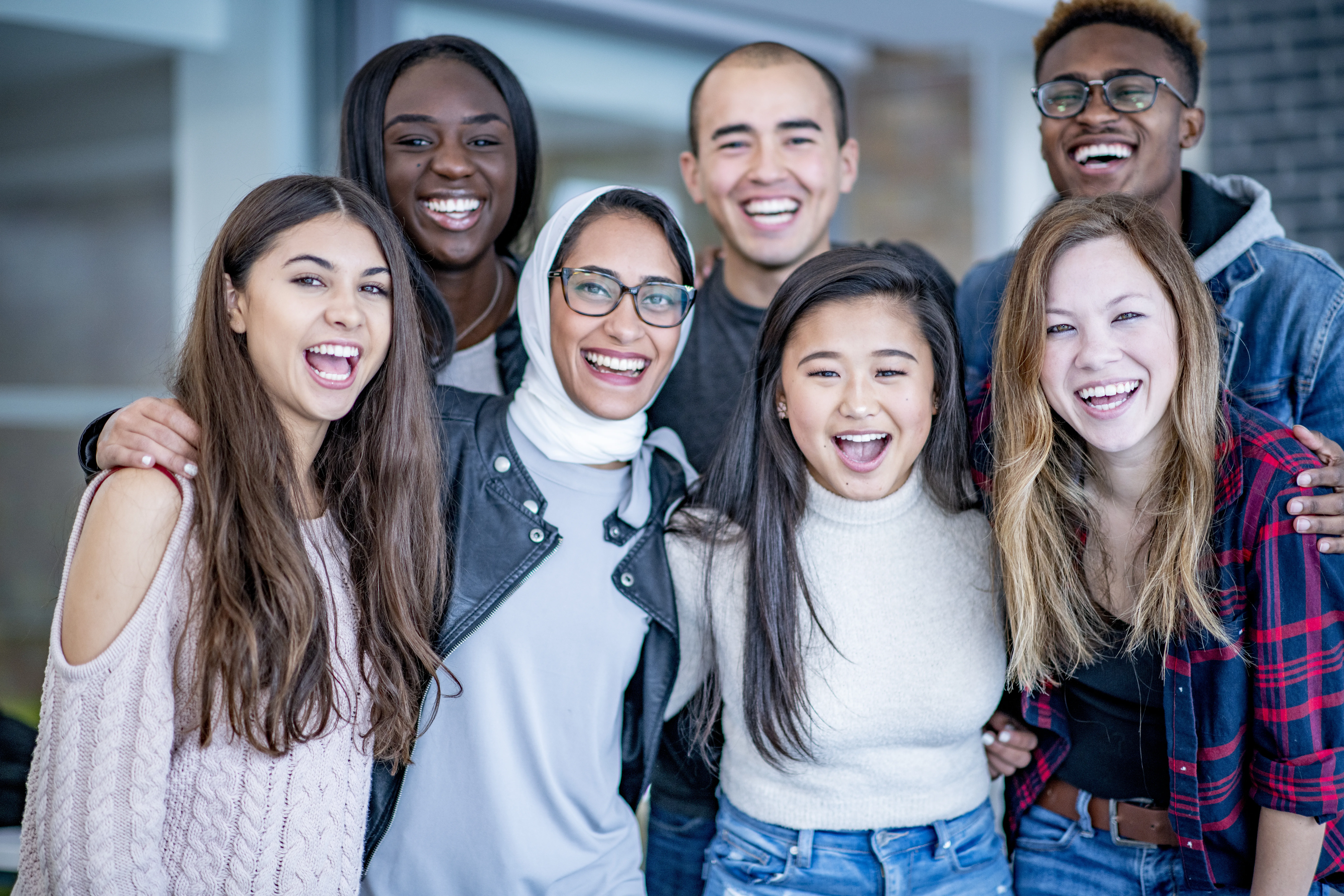 A varying group of young adults stand and smile with their arms around each other. They could be university or college students.
